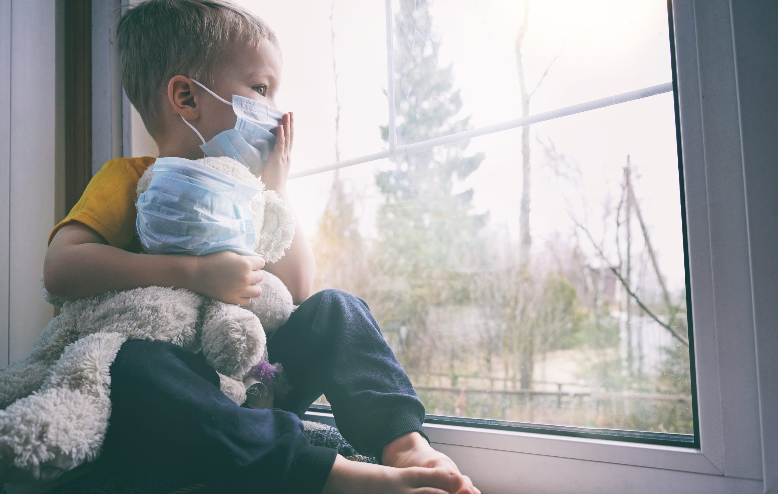 Child and his teddy bear wearing a mask looking out the window