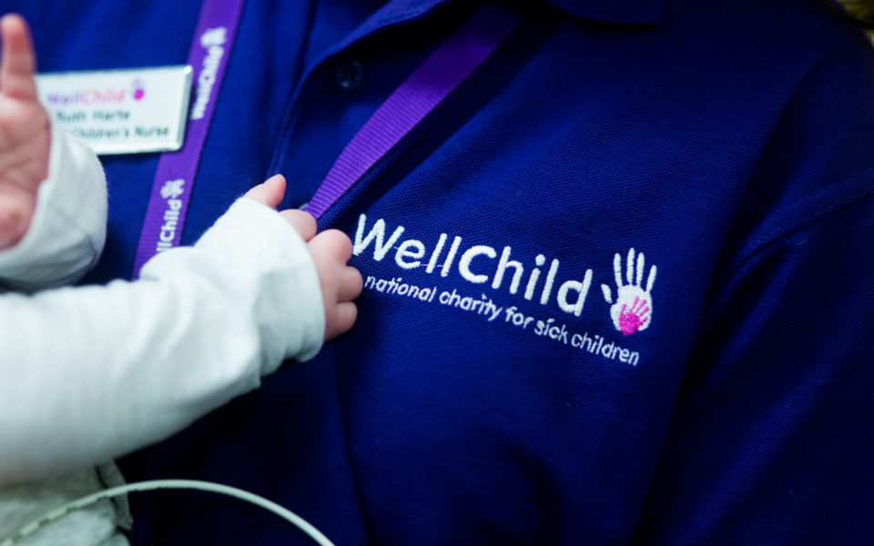 A baby being held by a WellChild Nurse. The baby's hand touching the WellChild logo on the Nurse's top.