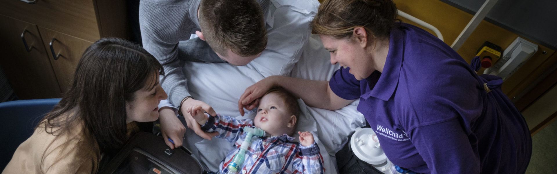 Ariel shot of a young boy lying in a hospital bed with his parents and WellChild Nurse looking over him.