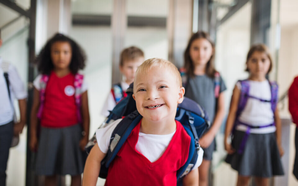 A down-syndrome school boy with bag and group of children in corridor, walking.